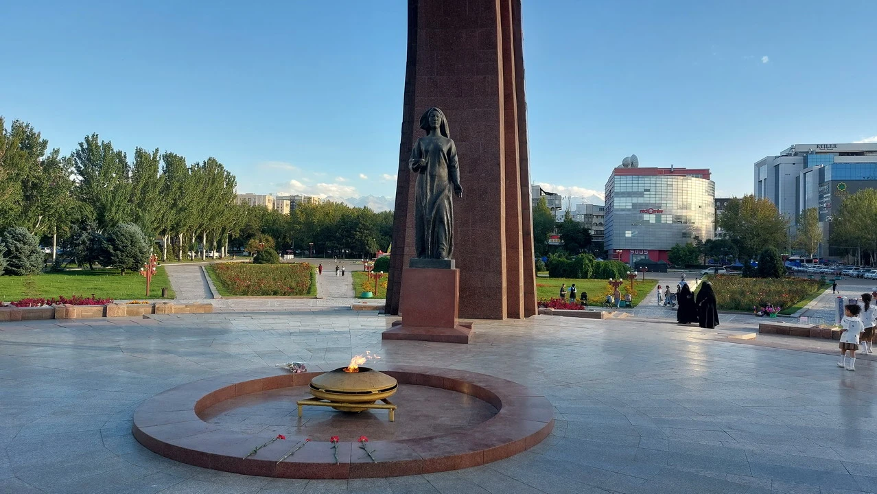 Una statua eretta si trova in una piazza aperta con alberi e edifici sullo sfondo.