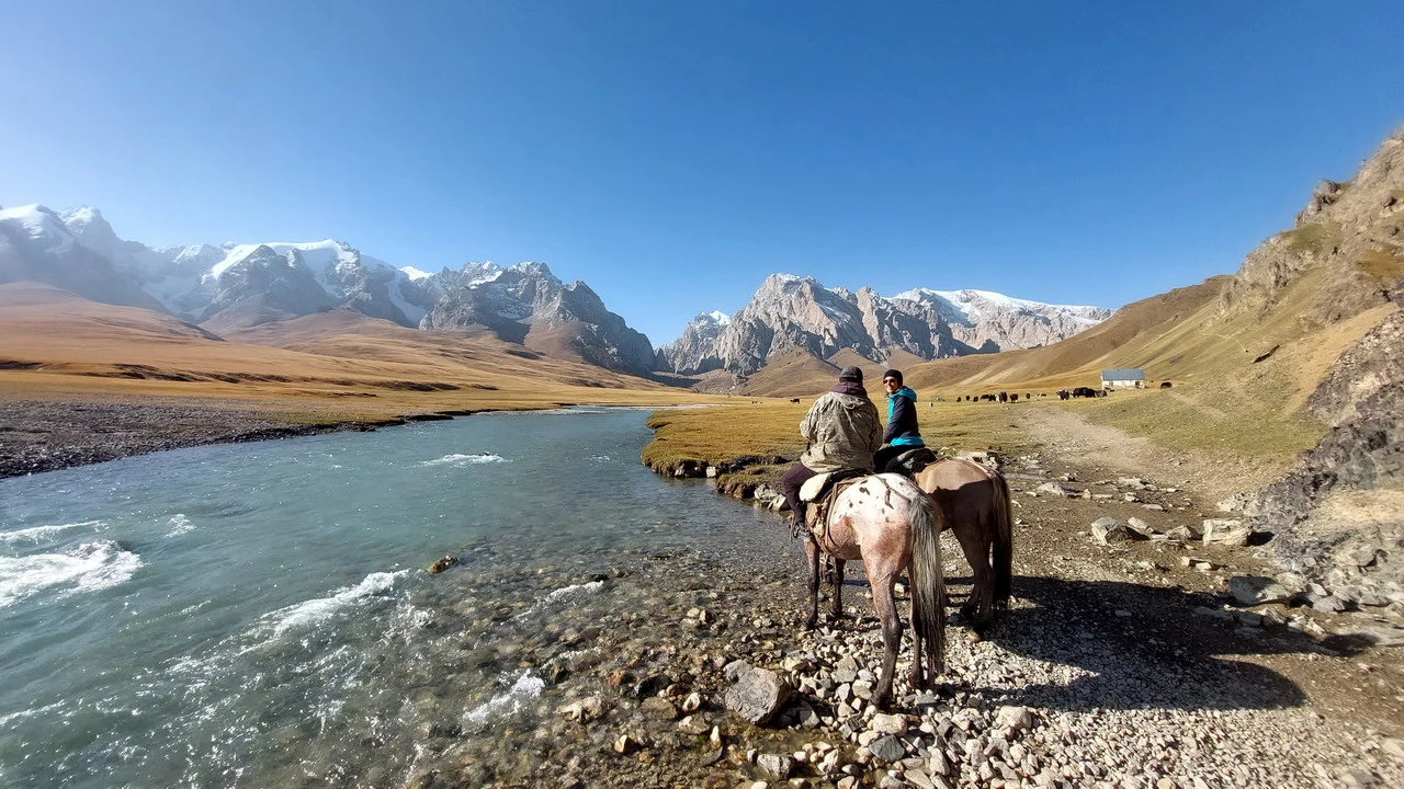 Due cavalieri siedono vicino a un fiume in una valle montuosa e arida.