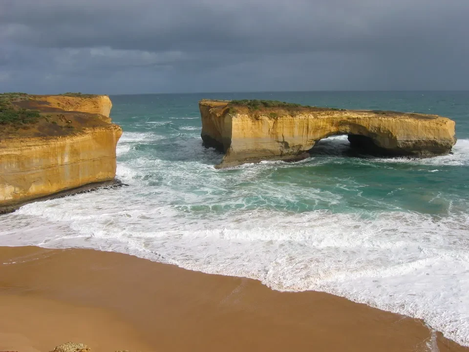 Formazioni rocciose costiere con un arco marino e una spiaggia sabbiosa sotto un cielo nuvoloso in Australia.