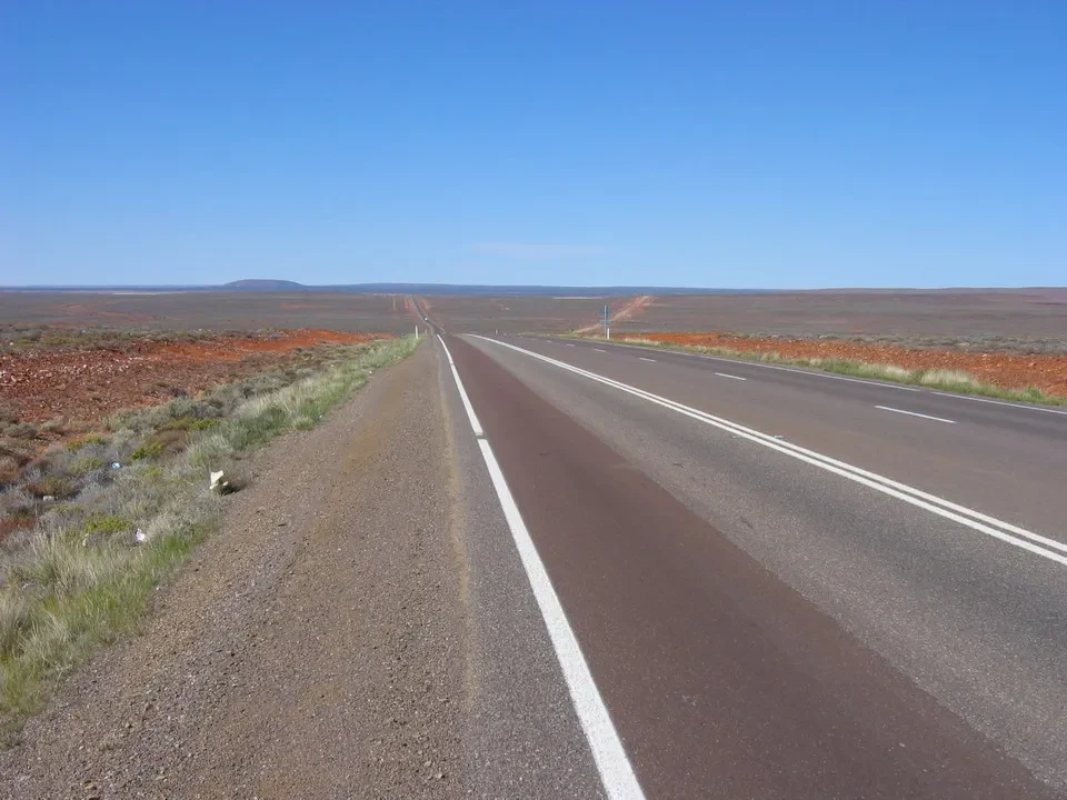 Una strada asfaltata si estende attraverso un paesaggio arido australiano sotto un cielo azzurro.