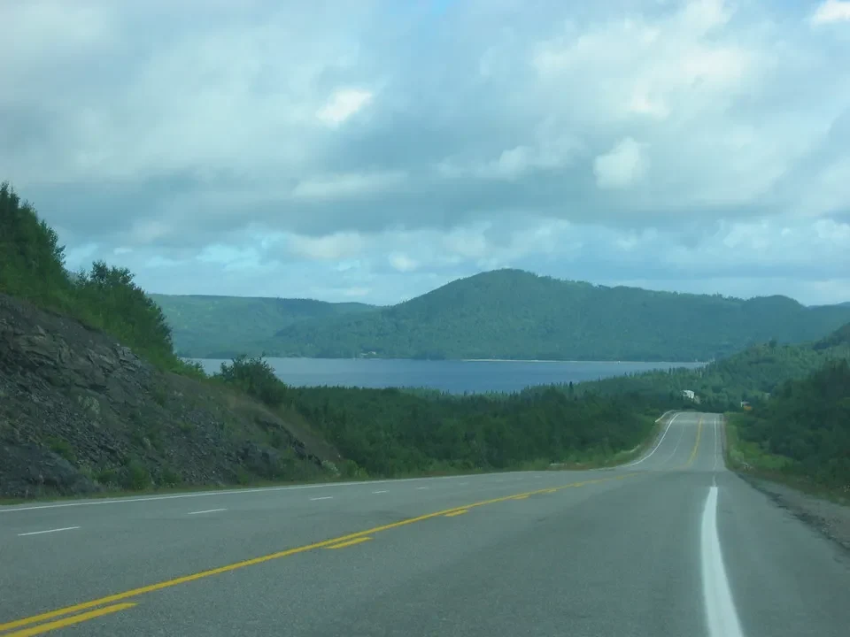 Una strada asfaltata attraversa un paesaggio lacustre e boscoso in Canada orientale sotto un cielo nuvoloso.
