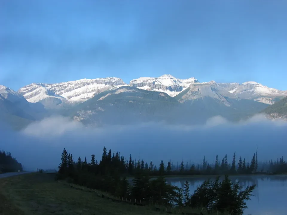 Montagne innevate parzialmente nascoste dalla nebbia mattutina sopra un lago e una foresta.
